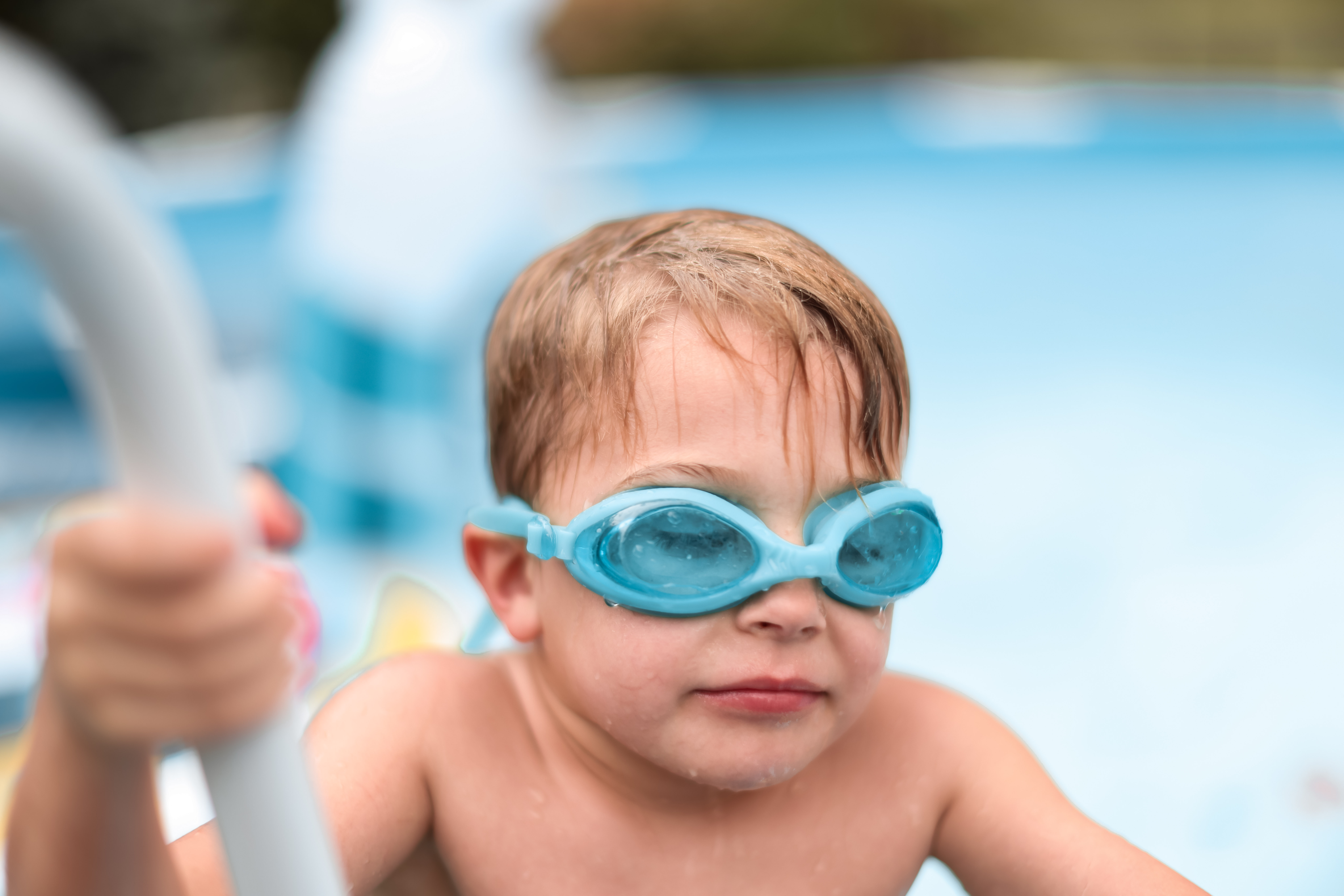 Young child wearing blue swim goggles in a pool, holding onto the pool ladder.