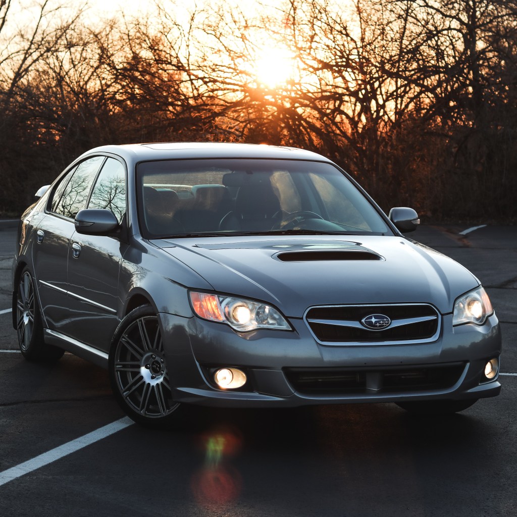 Silver Subaru Legacy sedan parked in a lot during sunset with trees silhouetted in the background.