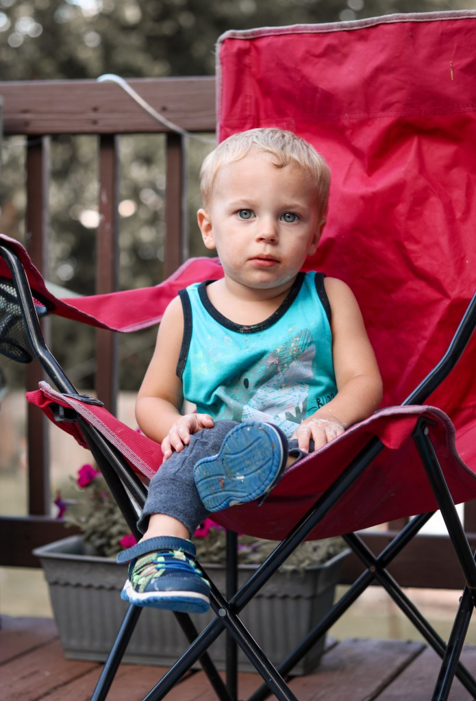 Toddler sitting in a large red camping chair on a wooden deck.