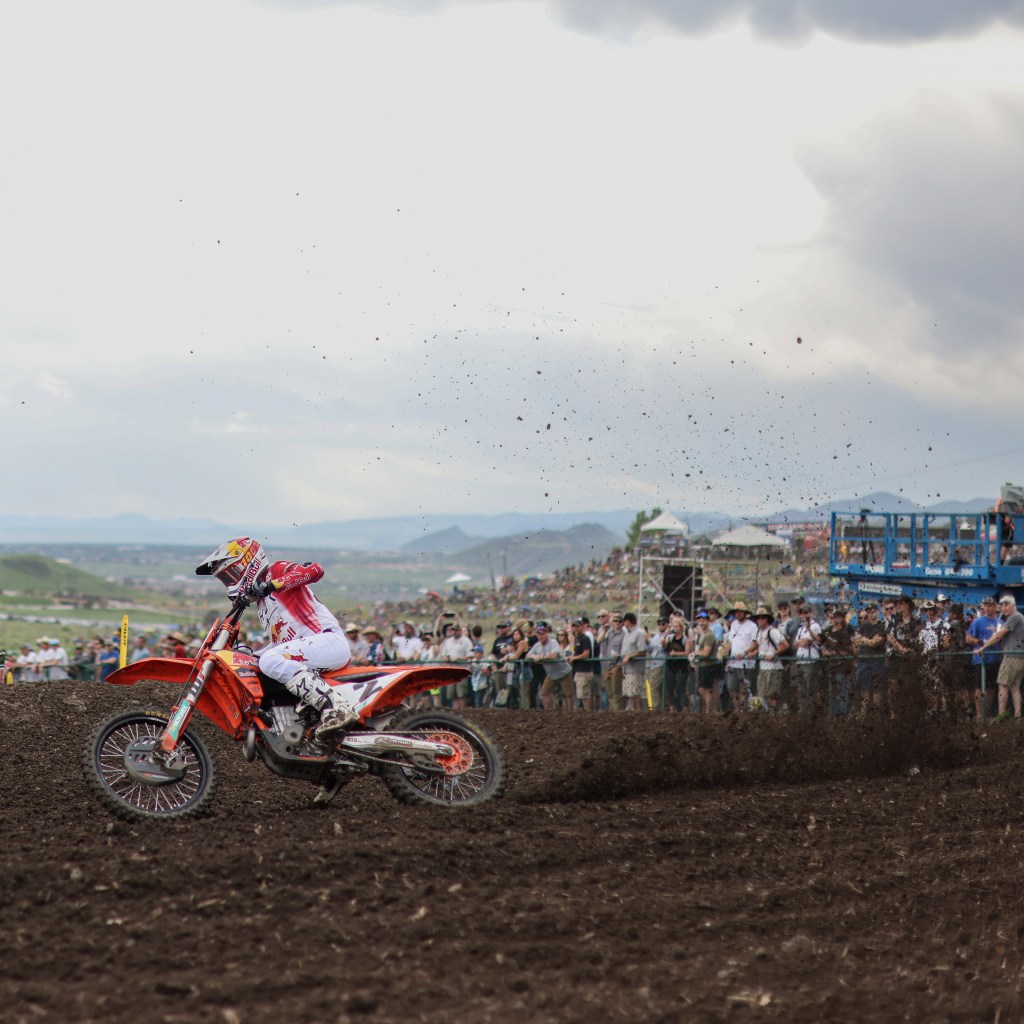 Cooper Webb (#2) on a KTM, kicking up roost while leaning into a turn at Thunder Valley, with mountains in the background and a crowd watching.