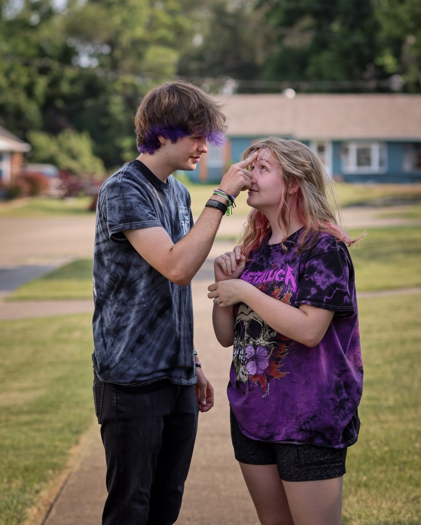 A young couple wearing matching acid-wash t-shirts share a playful moment on a neighborhood sidewalk. The boy gently pushes up the girl’s glasses in a goofy way while she gazes up into his eyes, smiling. Both have colorful hair, adding to their unique style.