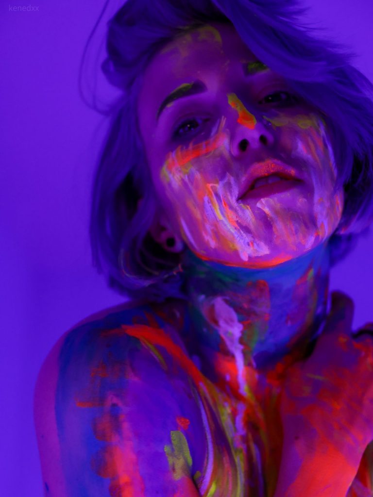Artistic self-portrait of a person covered in neon glow-in-the-dark paint, standing in front of a purple backdrop under black light. Short purple hair partially covers the face, and the person’s right hand holds their left shoulder. The lips are parted, and the head is tilted upwards while looking directly at the camera, showcasing a moody and dramatic vibe.