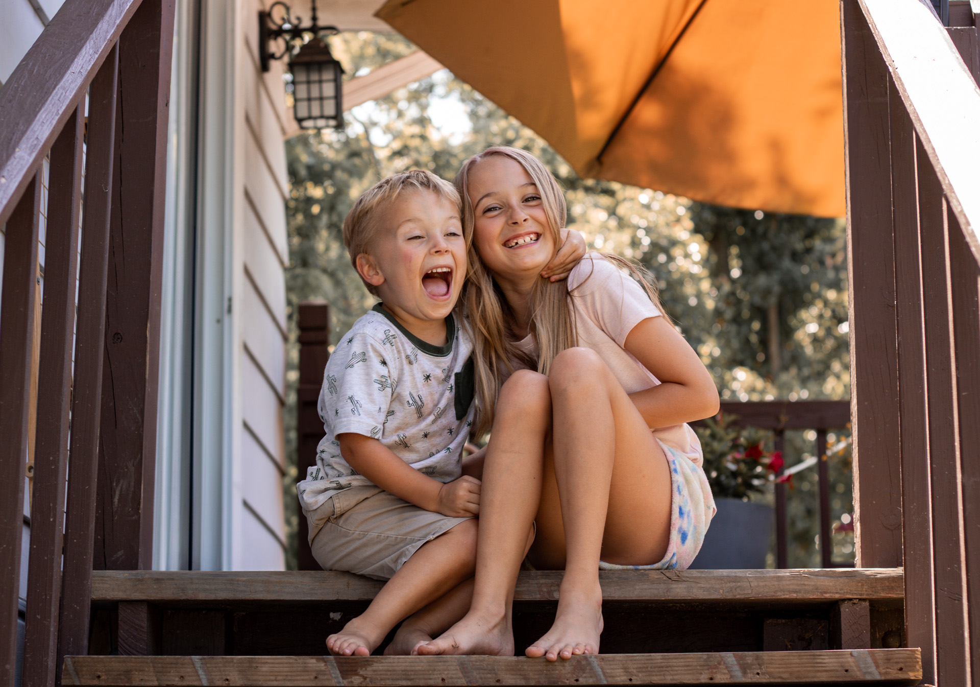Happy brother and sister laughing together on their mom's porch, with the brother playfully hugging the sister.