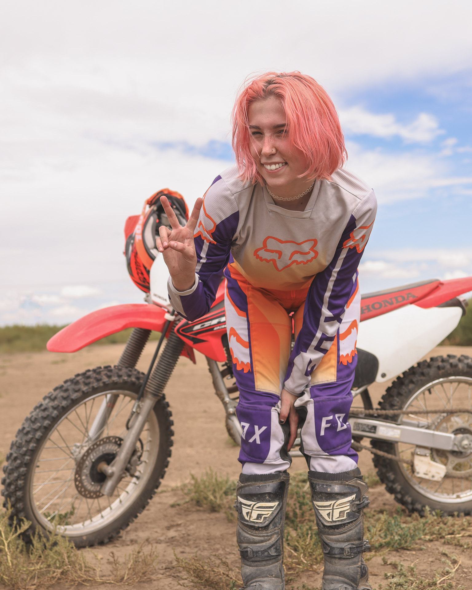 A self-portrait of a woman with short peach-colored hair standing in front of her Honda dirt bike in the desert. She is leaning toward the camera, smiling, and making a peace sign while wearing dirt bike gear in the summer sun.