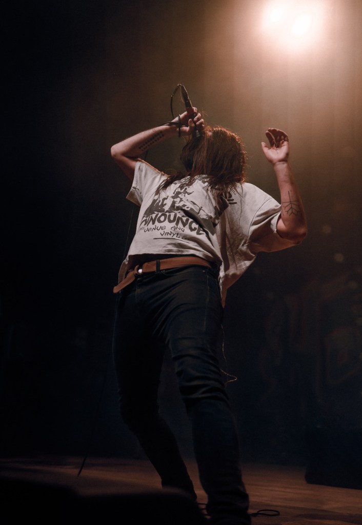 Nathan "Tades" Sanville, lead singer of Hot Mulligan, sings passionately on stage during the Choose Your Fight Tour. He holds the microphone close, his hair covering his eyes as he leans back, hands gripping the mic cord. The stage is dark and moody, highlighting the emotion in his performance.