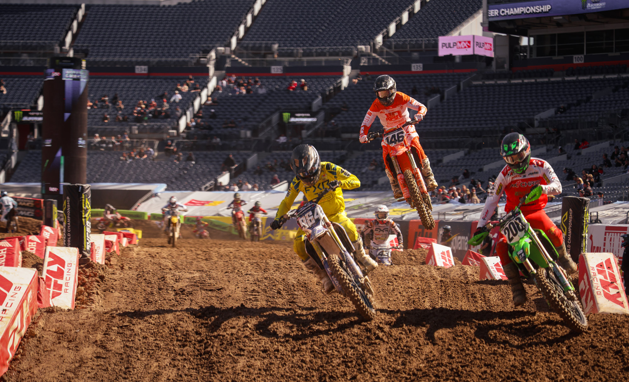 Three dirt bike riders in denver, colorado on media day for supercross 2025. Two leaning into a corner with one jumping between them.